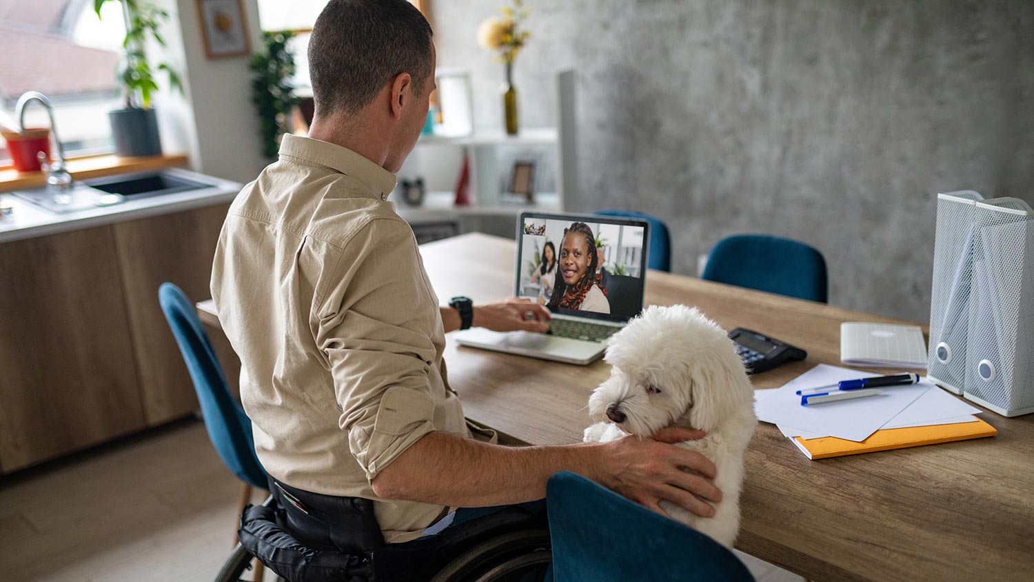 Man in a wheelchair on a laptop video call at a dining table while petting a small white dog