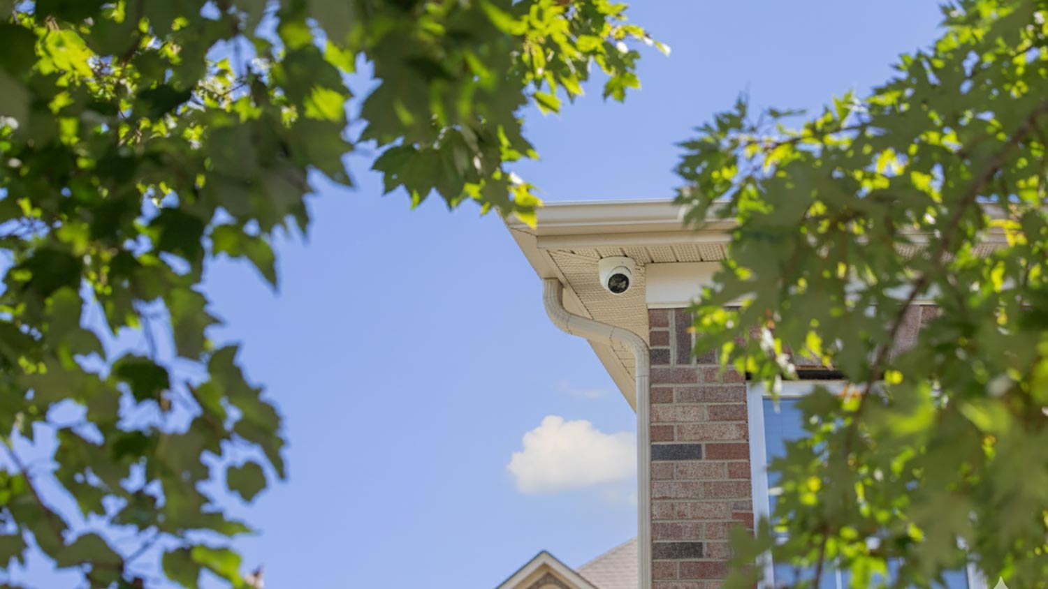 White dome security camera mounted under the roof eave of a brick house against a blue sky